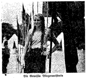Schwarz-Weiß-Foto in schlechter Qualität. Es zeigt ein Mädchen mit langen blonden Zöpfen und einer Hand an der Hüfte vor Fahnen.